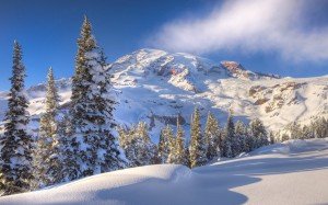 Berggipfelgrat, in Schnee gehüllt, davor unberührte Nadelbäume und reinweißer Schnee vor einem nebelwolkig-blauen Himmel