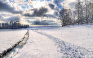 HDR-Foto eines schmalen Bachs, der sich schnurgerade neben einem Weg durch eine sonst unberührte Schneelandschaft zieht, rechts ein Waldrand, hinten ein anderer Wald mit einer Hütte. Himmel wolkig mit durchbrechenden Sonnenstrahlen