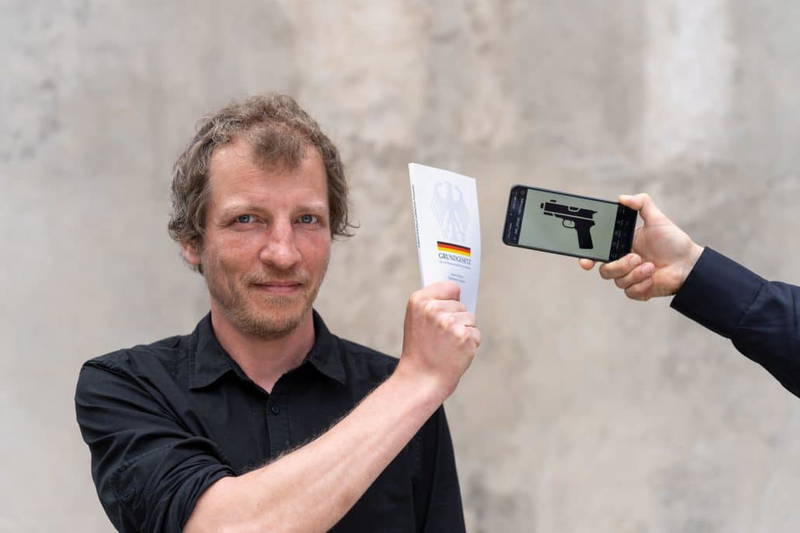 Mann zeigt Grundgesetz und Smartphone mit Waffe.